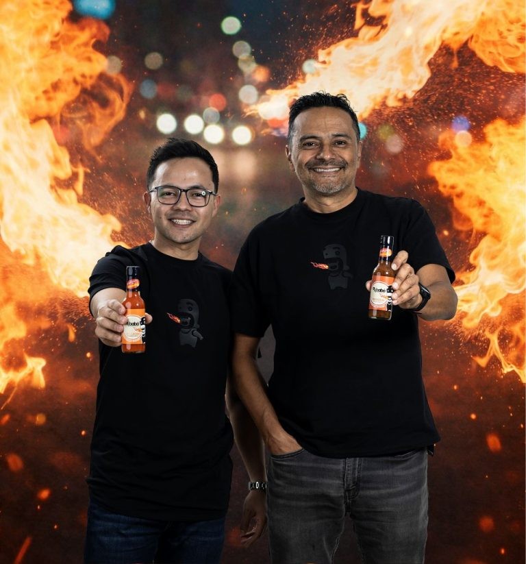 Padre e hijo, fundadores de Ajibaba, con sus camisetas y frascos de salsa frente a un fondo de fuego